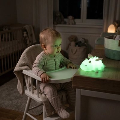 bebe assis dans une chaise haute observant une veilleuse bébé dinosaure eclairee en vert sur une table de la chambre